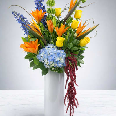Tall mixed bouquet in a white vase with orange lilies, yellow roses, and blue flowers