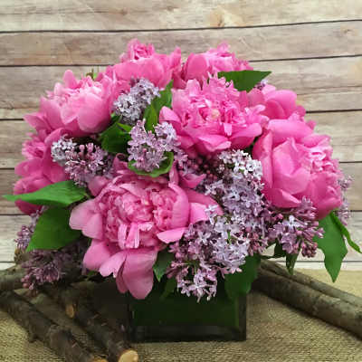 Pink peonies and lilac flowers arranged in a square glass vase