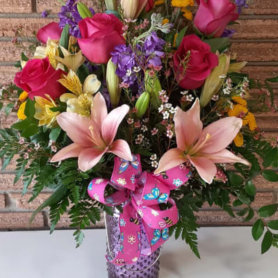 Mixed bouquet of pink roses, peach lilies, and yellow blooms in a purple vase with a pink butterfly ribbon