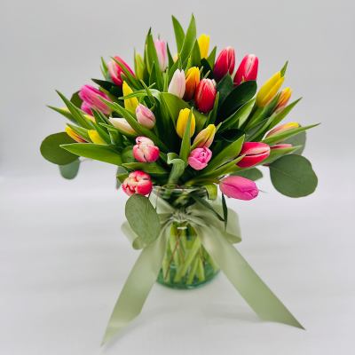 Mixed pink, yellow, and white tulips in a clear glass vase tied with a wide green ribbon