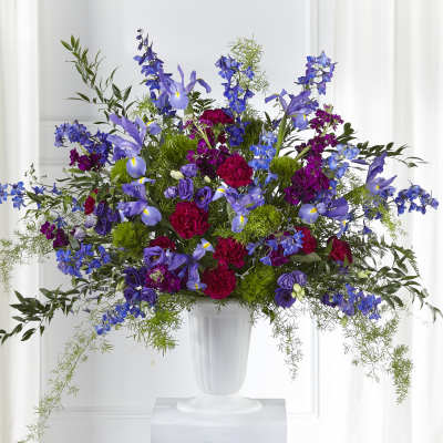 Tall arrangement of purple irises, blue delphinium, and red carnations in a white pedestal vase