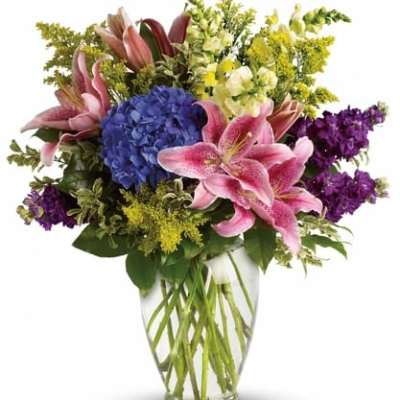 Mixed bouquet of pink lilies, blue hydrangea, and purple and yellow blooms in a clear glass vase