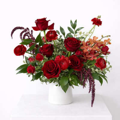 Arrangement of deep red roses, ranunculus, and orange orchids in a white vase
