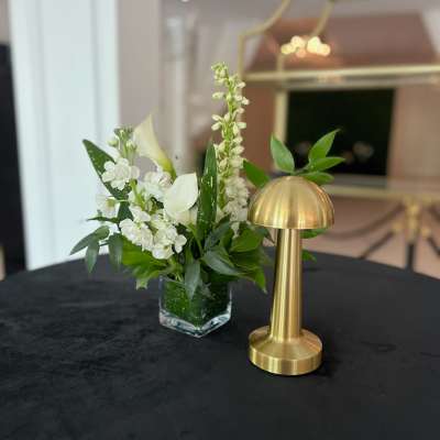 White floral arrangement in a clear square vase beside a gold table lamp