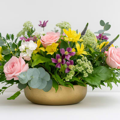Mixed bouquet of pink roses, yellow flowers, and purple blooms in a gold bowl