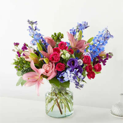 Mixed bouquet of pink lilies, roses, and blue delphinium in a clear glass vase