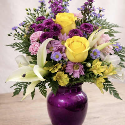 Mixed bouquet of yellow roses, purple daisies, and white lilies in a purple vase