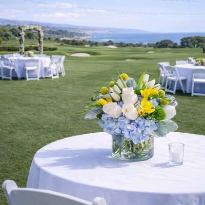 Low blue, white, and yellow flower centerpiece in a clear vase on an outdoor table with white linens