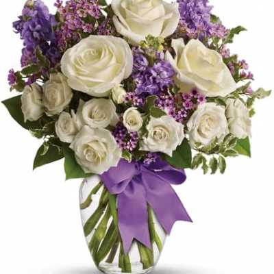 White roses and purple flowers arranged in a glass vase with a purple ribbon.
