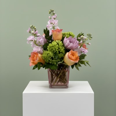 Mixed bouquet of peach roses, lavender blooms, and green flowers in a pink vase