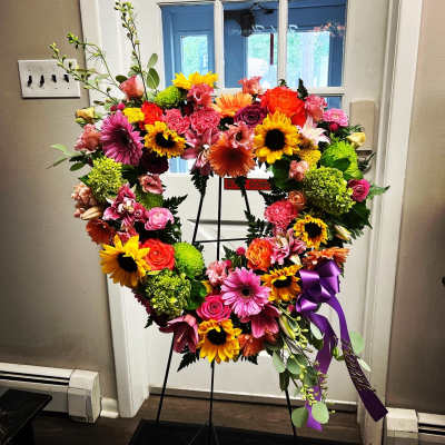 Heart-shaped floral wreath on a stand with bright mixed blooms and a purple ribbon