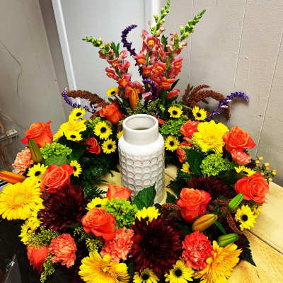 Circular floral arrangement with orange roses, yellow daisies, and a white vase in the center