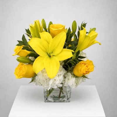 Yellow lilies and roses arranged in a clear square vase with white flowers below