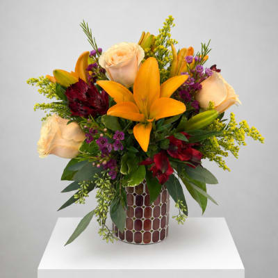 Mixed bouquet with orange lilies, cream roses, and purple accents in a patterned vase