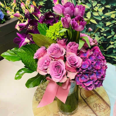 Pink roses and purple flowers arranged in a glass vase with a pink ribbon.