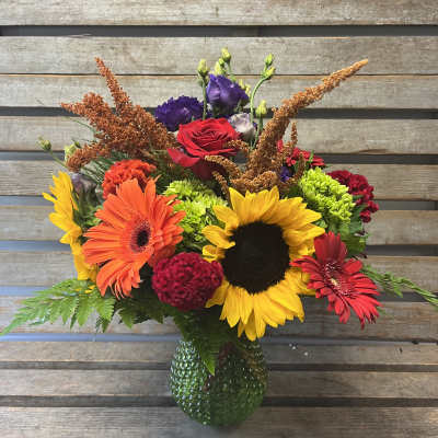 Colorful mixed bouquet with sunflowers, gerbera daisies, and roses in a green glass vase