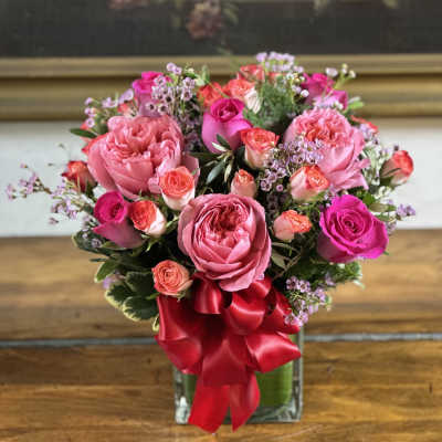 Compact arrangement of pink and hot pink roses in a clear glass vase with a red ribbon bow.