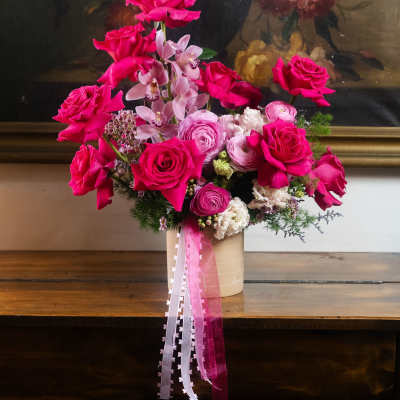 Hot pink roses and pale pink orchids in a beige vase with ribbon tails