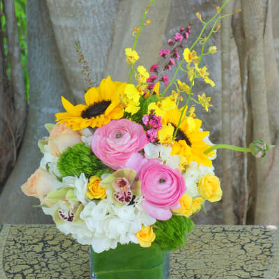 Bright mixed bouquet with sunflowers, pink ranunculus, orchids, and hydrangeas in a glass cube vase