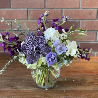Low glass vase with purple orchid, lavender lisianthus, white roses and mixed blooms on a table.