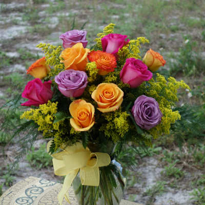 Mixed bouquet of yellow, orange, pink, and lavender roses in a clear vase with a yellow bow