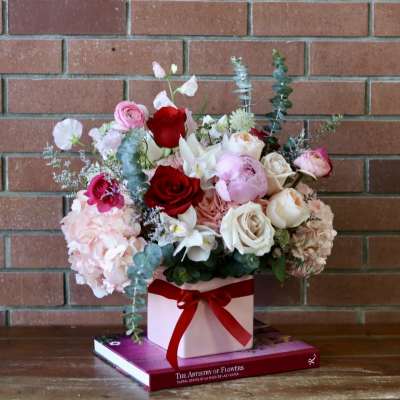 Mixed bouquet of roses, peonies, and hydrangeas in a pink box with a red ribbon