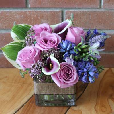 Short arrangement of pink roses, purple calla lilies, and blue flowers in a square glass vase
