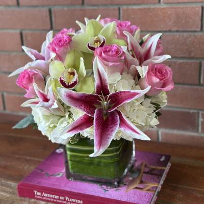 Compact arrangement of pink lilies, roses, green orchids, and white hydrangeas in a square glass vase.