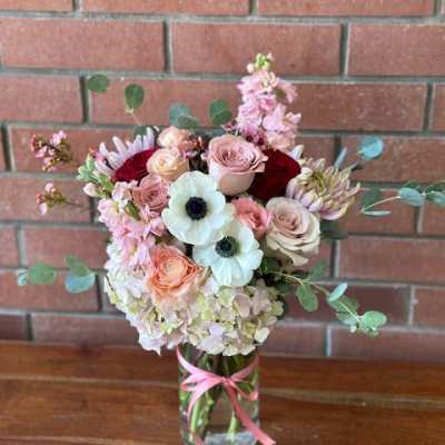 Tall mixed bouquet of soft pink, white, and peach flowers in a clear vase with a pink ribbon.