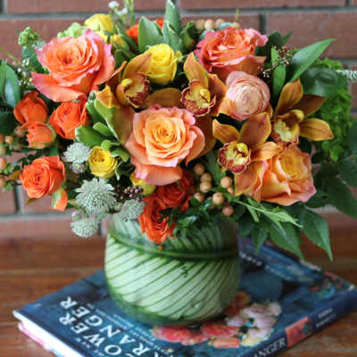 Low arrangement of orange and yellow roses with orchids in a round glass vase wrapped with green leaves