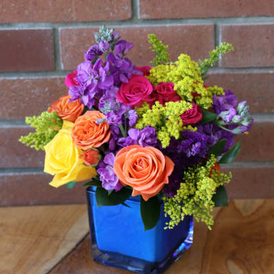 Bright mixed bouquet of orange and yellow roses with purple flowers in a blue glass cube vase