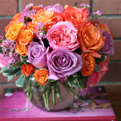 Round arrangement of orange, pink, and lavender roses in a clear glass bowl vase