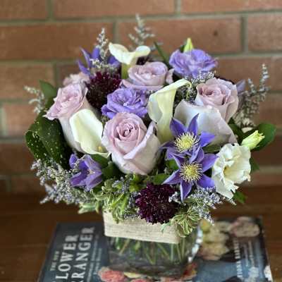 Low lavender and white flower arrangement with roses and calla lilies in a glass cube vase on top of books