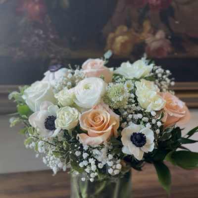 Round arrangement of white and peach roses, ranunculus, and anemones in a clear glass vase.