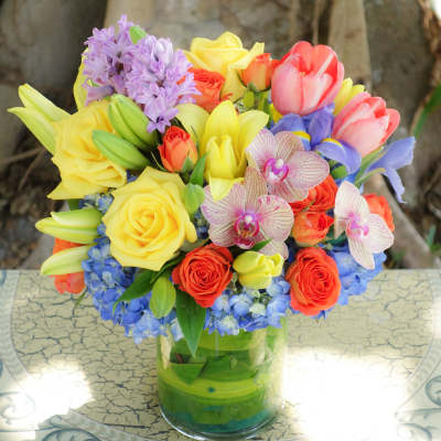 Bright mixed bouquet of yellow roses and lilies with tulips, orchids, and blue hydrangea in a clear glass vase