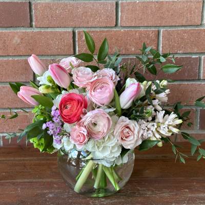 Low glass vase of pink tulips, ranunculus, roses, and white blooms in a rounded bouquet.