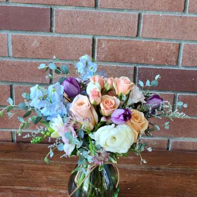 Peach roses, white ranunculus, and purple tulips arranged in a round green glass vase.
