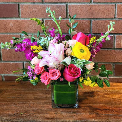 Bright mixed bouquet of pink roses and yellow ranunculus in a square glass vase lined with green leaves