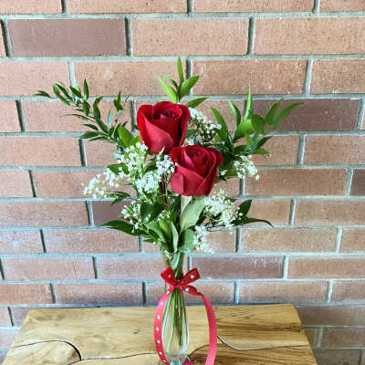 Two red roses in a clear glass vase with white filler flowers and a red ribbon