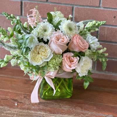 Compact arrangement of blush roses and white blooms in a green glass cube vase with pink ribbon