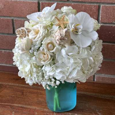 White hydrangeas, roses, and orchids with seashells in a blue glass vase