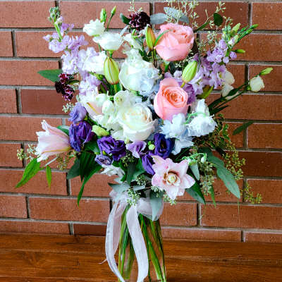 Tall bouquet of pink roses, purple lisianthus, and lilies in a clear glass vase with ribbon