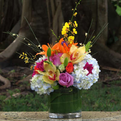 Low glass vase of colorful orchids, lilies, roses, and hydrangeas arranged in a compact mixed bouquet.