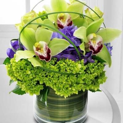 Compact arrangement of green cymbidium orchids and purple flowers in a clear glass cylinder vase