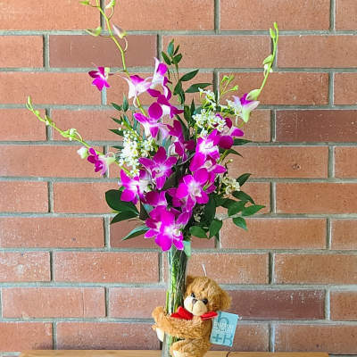 Tall vase of purple and white orchids with white filler flowers and a small teddy bear hugging the vase