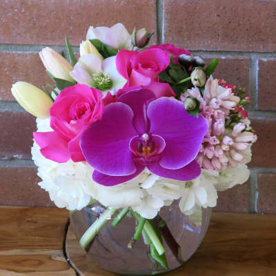 Compact bouquet of pink orchids, roses, tulips, and white hydrangeas in a round clear glass vase