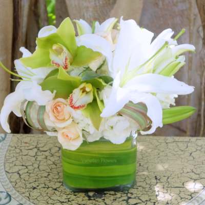 Low arrangement of white lilies, green orchids, and peach roses in a square glass vase
