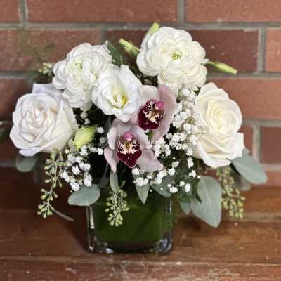 Compact arrangement of white roses and ranunculus with pink orchids in a square glass vase