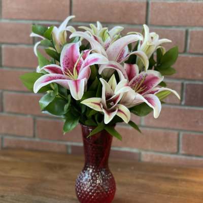 Pink and white lilies arranged in a red textured glass vase on a wooden surface.