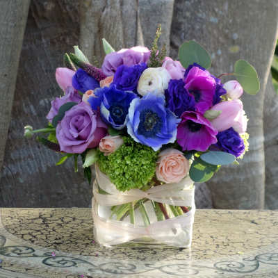 Compact arrangement of purple roses, blue and fuchsia anemones, and peach blooms in a square glass vase.